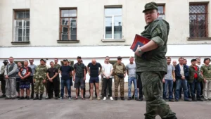 Офіцер у камуфляжі з папками проходить повз шеренгу чоловіків біля будівлі